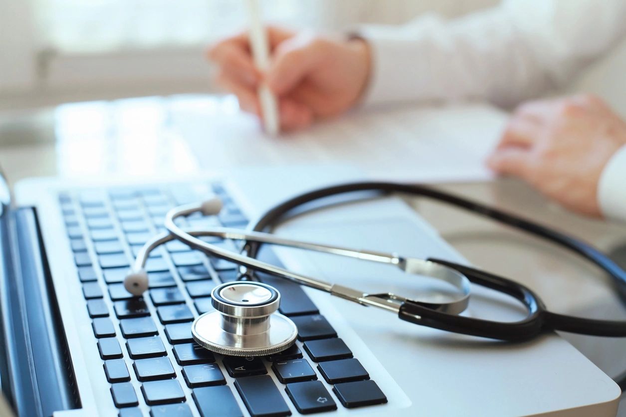 A doctor filling out paperwork with a stethoscope on a laptop